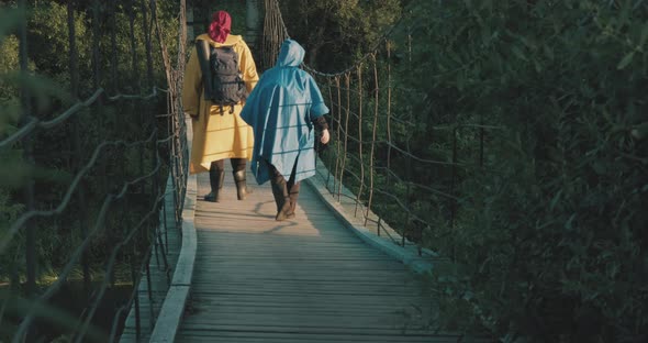 Man and Woman Hikers in Raincoats Walk on Suspension Bridge