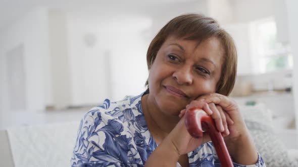 Happy african american senior woman sitting leaning on walking stick, looking away smiling alt