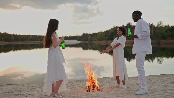 Group of Friends Having Fun on a Lake Beach Drinking Near Campfire