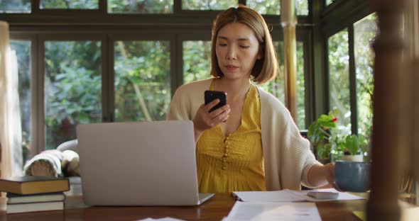 Asian woman sitting at table working from home and using smartphone alt