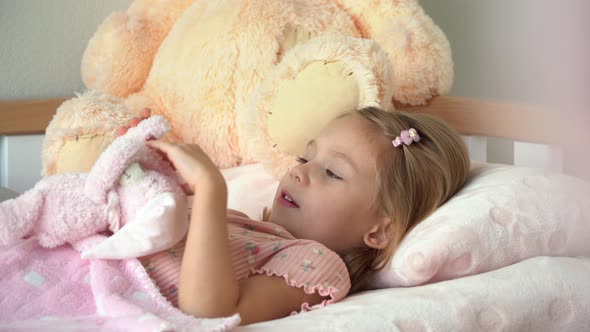 Little Girl Lies in Bed Before Bedtime and Plays with Toy