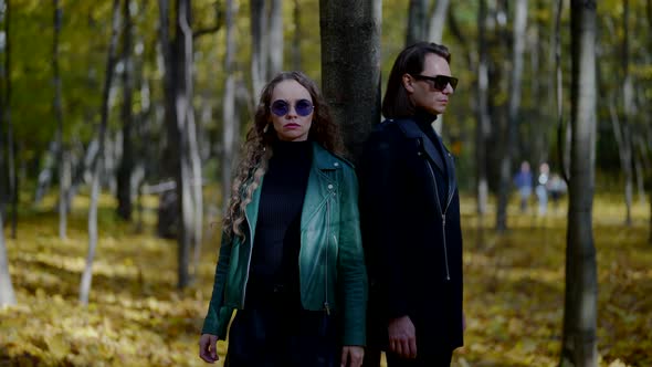 a Man and a Blonde in Leather Jackets and Sunglasses Stand with Their Backs to a Tree in an Autumn alt