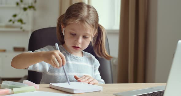 Portrait of Little Cute Preschool Girl Wears Headphones Sits at Home Table Alone Looks at Laptop alt