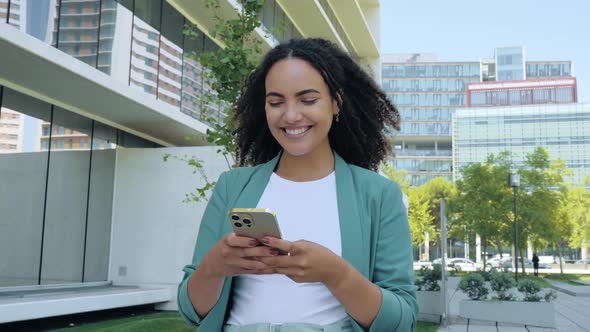 Positive Friendly Pretty Mixed Race Young Woman with Curly Hair Stylishly Dressed Using Smartphone alt