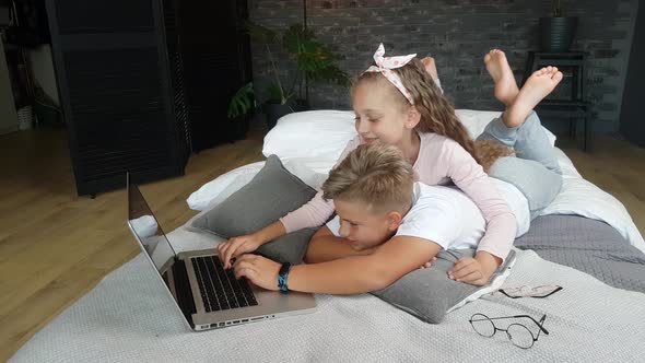 Children - a girl and a boy in home clothes lie on the bed embracing and play in a laptop alt