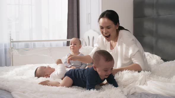 Loving Asian Mom Sitting on Bed with 1-Year-Old Triplets alt