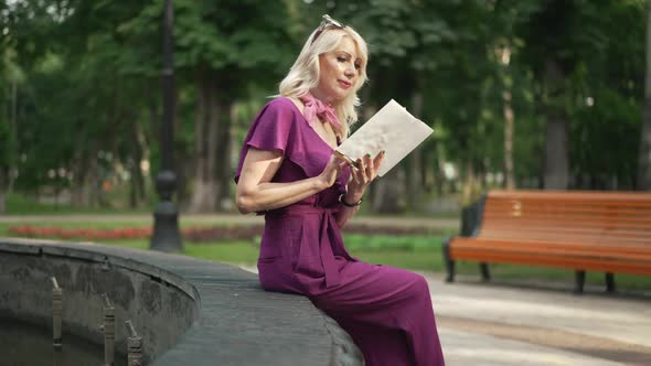 Side View Absorbed Female Reader Sitting at Fountain in City Park Turning Book Pages in Slow Motion alt