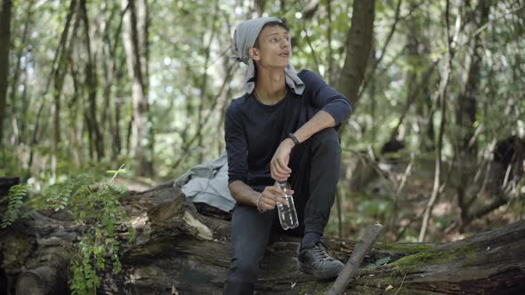 Nervous Scared Gypsy or Refugee Sitting on Log in Forest and Drinking Refreshing Water. Portrait of alt