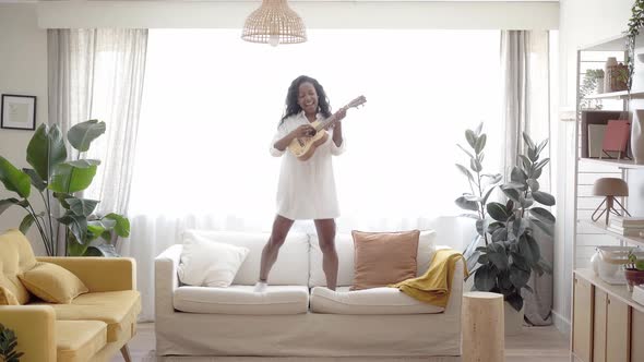 Motivated Woman Playing Ukelele Standing and Jumping on the Sofa Living Room alt