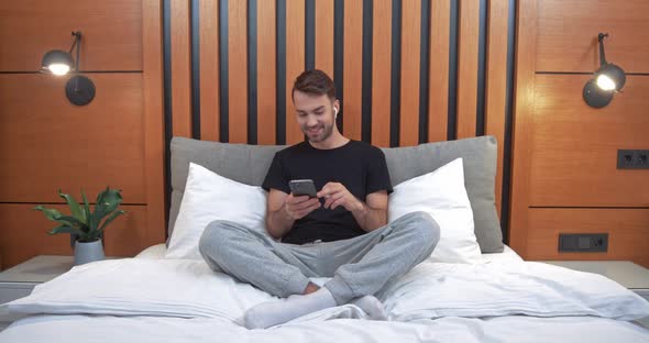 Young Happy Man Sitting on the Bed in the Bedroom and Listening To Music in Wireless Headphones alt