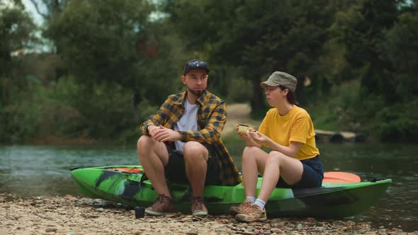 Picnic and kayaking. A young couple is sitting on a kayak, and they are eating lunch together. alt