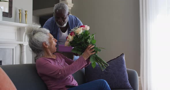 African american senior man giving gift box and flower bouquet to his wife at home alt