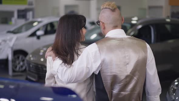Back View of Husband and Wife Hugging Discussing Car Purchase in Dealership in Slow Motion alt