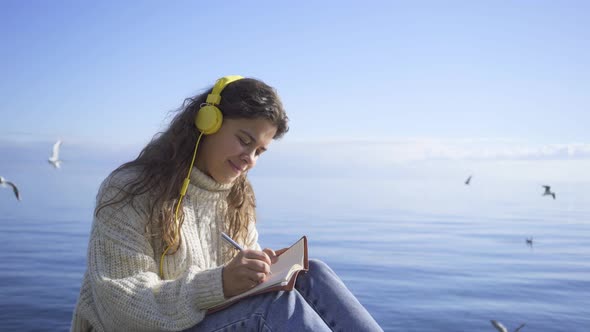 Positive Woman with Headphones Writes in Notebook Near Ocean