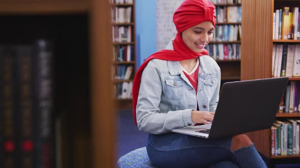 An Asian female student wearing a red hijab studying in a library and using laptop alt