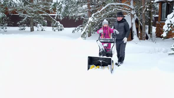 Father teaches his daughter in yard of house to remove snow with snowplow.