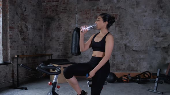Young Athletic Indian Asian Woman Sitting on a Bicycle and Drinking water from a Bottle alt