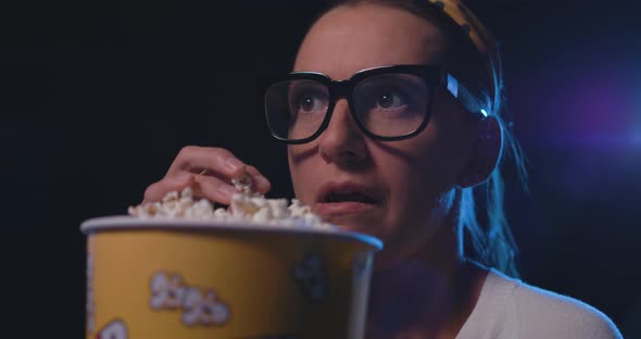Woman with glasses watching a scary horror movie at the cinema and eating popcorn