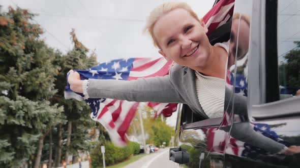 Happy Middle-aged Woman with an American Flag. Looks Out the Window of a Moving Car alt