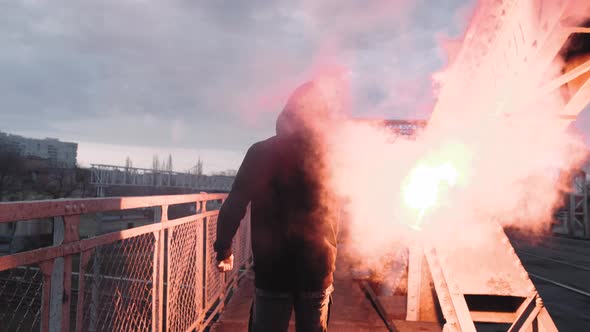 Back View of a Young Man in Hoodie and Balaclava with Red Burning Signal Flare on the Road Under an alt