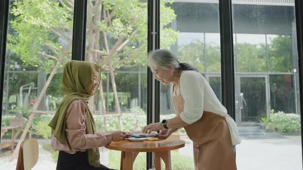 An elderly woman is serving coffee to a Muslim woman in a coffee shop.