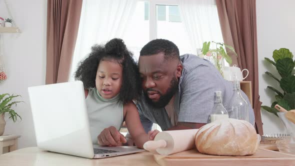 Cheerful African American dad teaching her daughter to cook by following method online from laptop