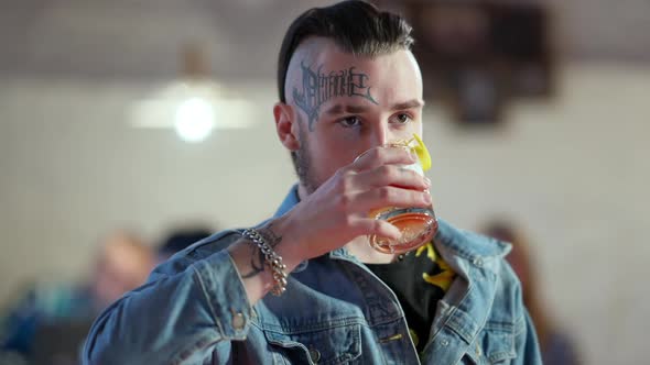 Handsome Confident Young Man with Tattoo on Face and Piercing Drinking Cocktail in Slow Motion alt