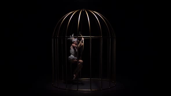Gymnast in Bird Costume Riding a Hoop in a Cage on Dark Stage. Black Background. Slow Motion alt