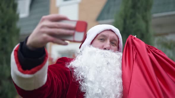 Caucasian Young Man in Red Santa Clause Costume Taking Selfie on Smartphone Standing Outdoors alt