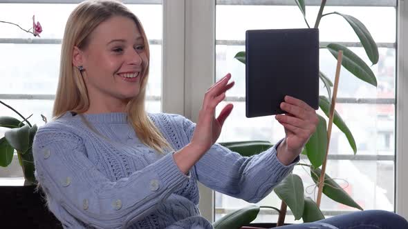 A Young Beautiful Woman Sits on a Chair By a Window and Has a Video Call on a Tablet alt
