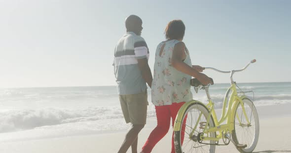 Senior african american couple walking with bicycle on sunny beach alt