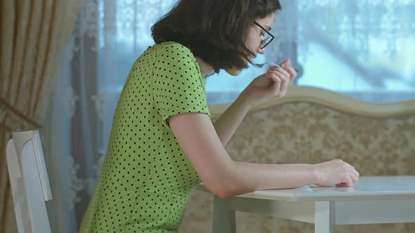 Brunette Girl in a Green Dress is Having Breakfast While Sitting at a Table at Home alt