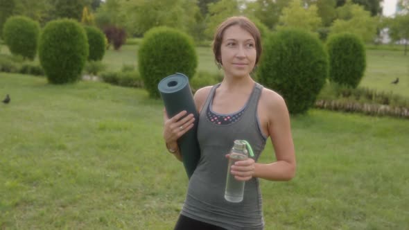 Portrait of a Young Woman Yoga Instructor with a Fitness Mat and a Bottle Against a Green Lawn