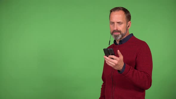 A Middleaged Caucasian Man Listens to Music with Earphones on and a Smartphone  Green Screen