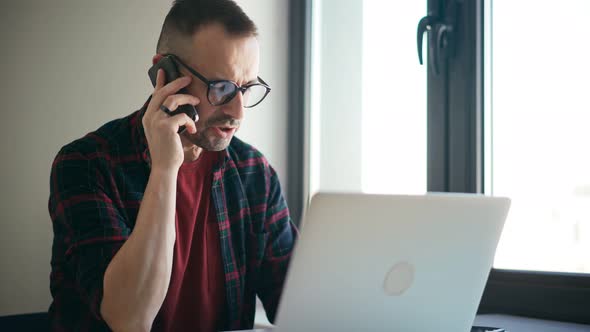 A Handsome Serious Man Is Talking on the Phone While Working on a Laptop at Home