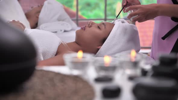 Beautician use cream mask on face of young Asian woman with brush in spa salon. alt