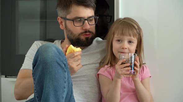 Father and Little Daughter Sit on Floor at Home Eat Ripe Apples and Drink Juice alt