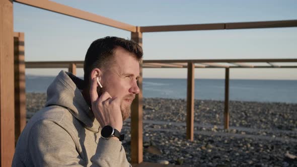 Man Is Checking His Handsfree Set in His Ear Sitting on Sea Shore alt