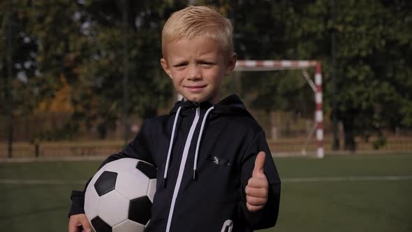 A Little Boy Player Sits on a Soccer Field with a Ball and Gives a Thumbs Up alt