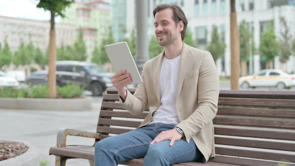 Online Video Chat on Tablet By Young Man Sitting on Bench alt