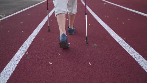 Closeup of a Senior Woman Legs Walking on a Stadium Track with Nordic Walking Poles Rear View alt
