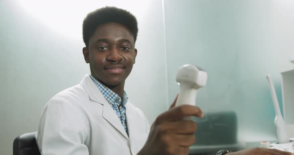 Smiling African Man Doctor with Ultrasound Transducer in Hand Working on Modern alt