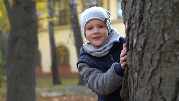 Beautiful Little Boy Hiding Behind Tree in Autumn Park alt