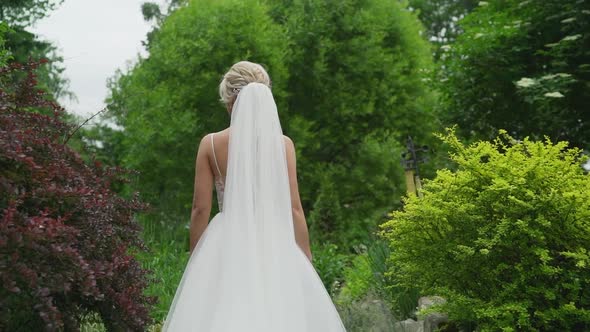 A Young Blonde in an Elegant Long White Wedding Dress with a Long Train Stands with Her Back to the alt