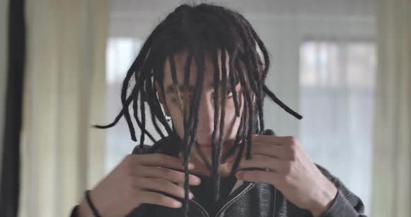Close-up Portrait of Serious Caucasian Young Man Taking Off Hood and Collecting Dreadlocks alt
