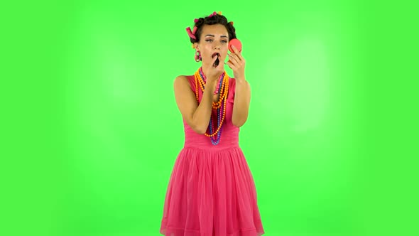 Girl with Curlers on Her Head in a Pink Dress Paints Her Lips Looking in Red Mirror. Green Screen alt