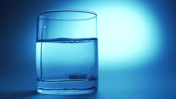 Pill Falling Down in Glass with Water. Effervescent Tablet Aspirin. Closeup on Blue Background alt