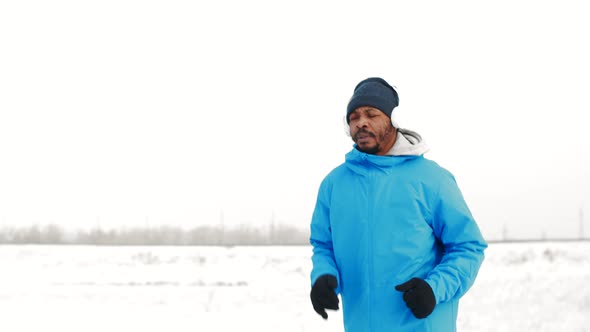 Black man at jacket, hat and headphones is jogging in winter outdoors, front view. African-American alt