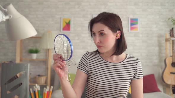 Distressed Young Hard of Hearing Woman Puts a Hearing Aid on Her Ear and Looks in the Mirror alt