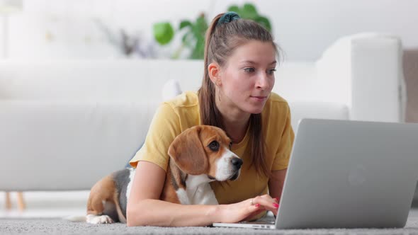 Woman Student Lying on Floor with Laptop and Doing Typing Homework Task with Pet Dog Spbi alt